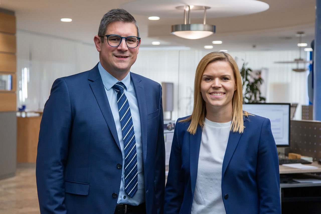 Vorstand, bestehend aus einem Mann und einer Frau stehen in Schalterhalle.