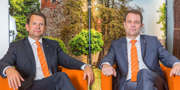 Zwei Bankvertreter mit orangefarbenen Krawatten sitzen auf orangefarbenen Foutons mit einer Parktapete im Hintergrund