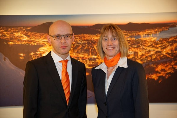 Ein Mann und eine Frau in schwarzen Anzügen mit orangefarbenen Krawatten und Schals vor einer Stadtlandschaft im Hintergrund