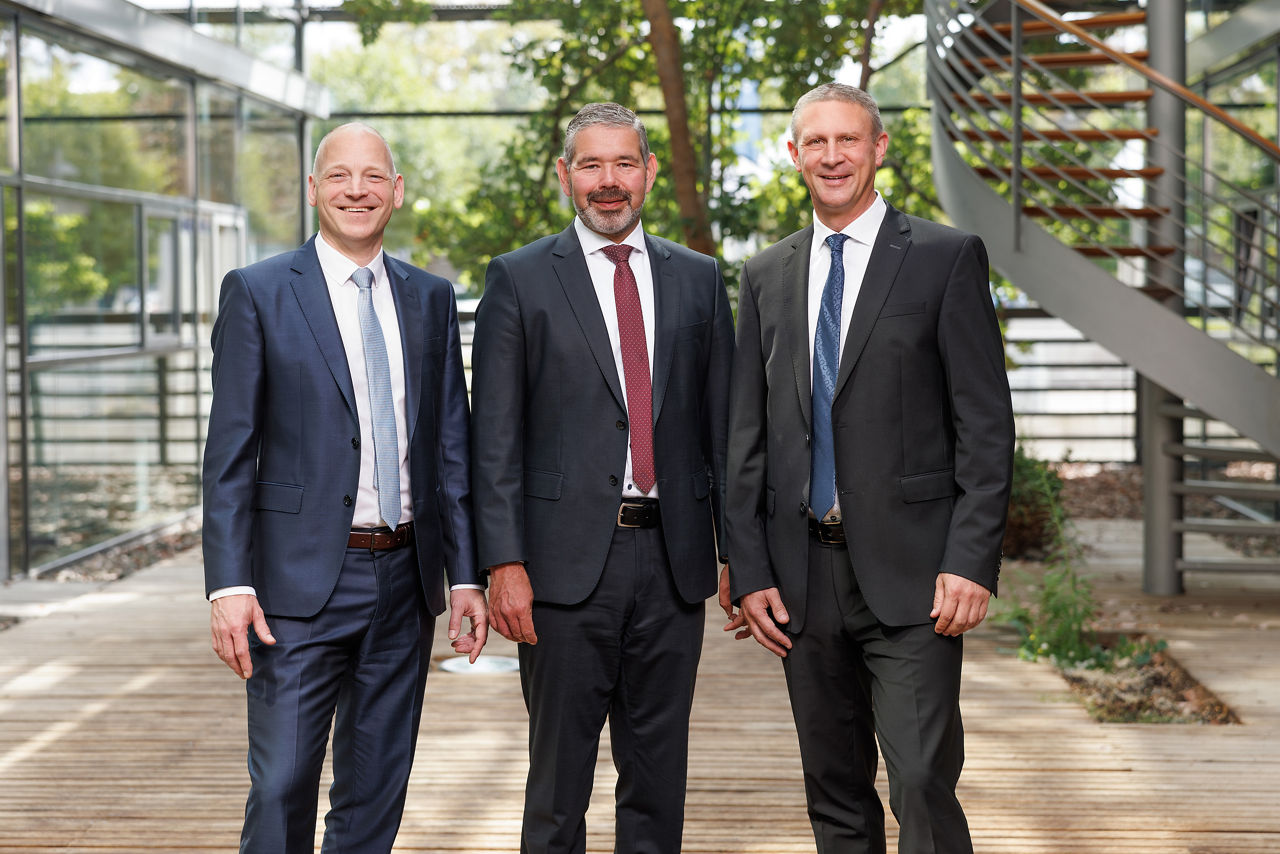 Drei Vorstandsmitglieder im Anzug stehend im Atrium