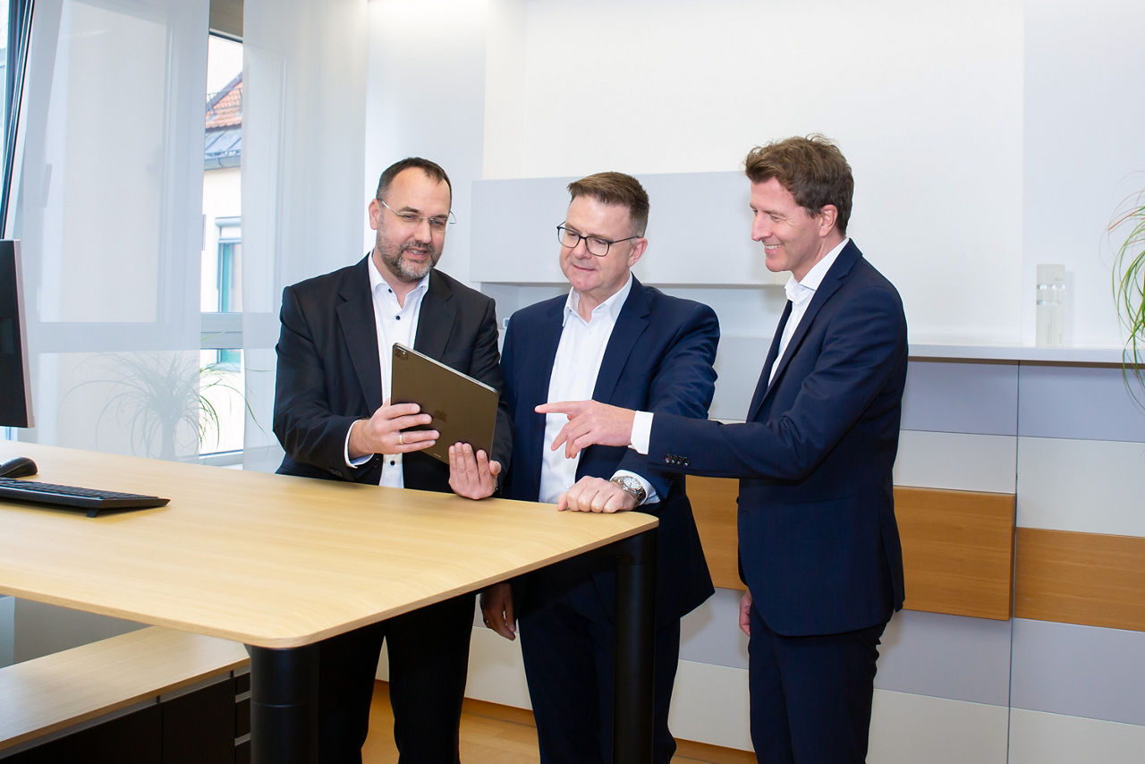 Drei Männer stehen hinter einem Schreibtisch und schauen in ein Tablet