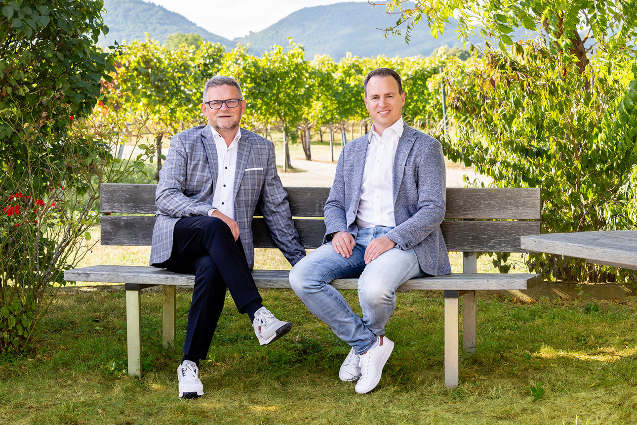 Zwei Personen sitzen auf einer Holzbank vor Weinreben in einer sonnigen Landschaft.