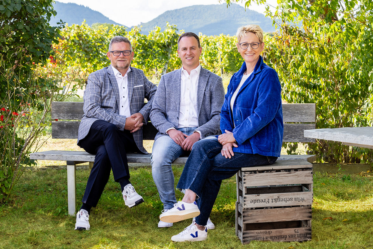 Drei Personen sitzen entspannt auf einer Bank im Grünen, mit Weinbergen und Bergen im Hintergrund.