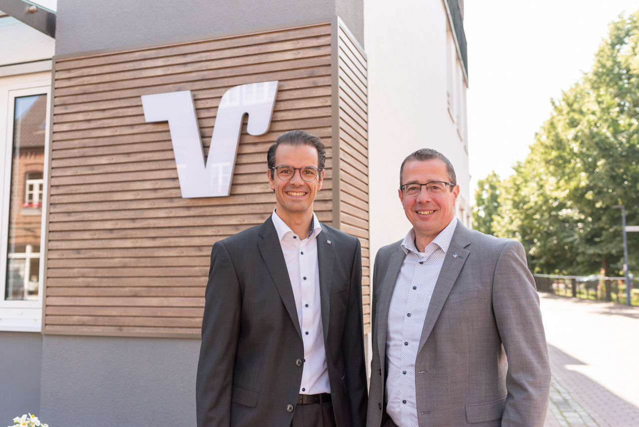 Die beiden Herren des Vorstandes stehen vor dem Bankgebäude vor einer Holzwand mit einem weißen Volksbankzeichen. 