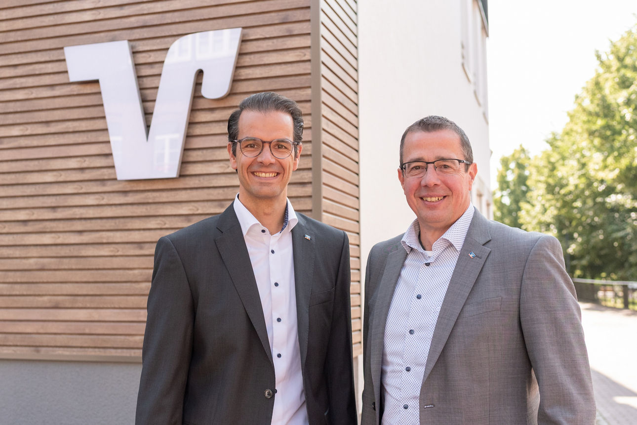 Die beiden Herren des Vorstandes stehen vor dem Bankgebäude vor einer Holzwand mit einem weißen Volksbankzeichen. 