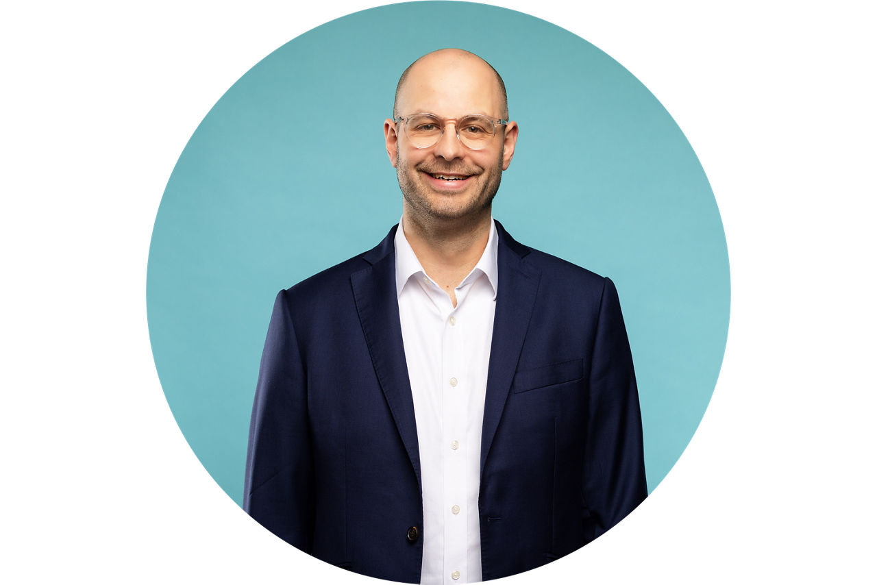 Porträt von Sebastian Emmert, Vorstand der VR Bank Fulda eG, vor hellblauem Hintergrund in dunklem Anzug und weißem Hemd.