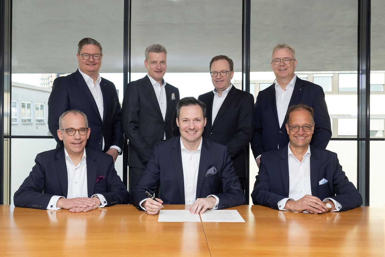 7 Herrn im Anzug sitzen und stehen vor eiem Tisch und unterschreiben eine Fussion der Volksbank