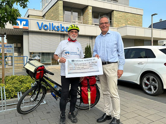 Ein alter, aktiver Mann mit Fahrradausrüstung, der mit einem Bankvertreter einen großen Scheck hält