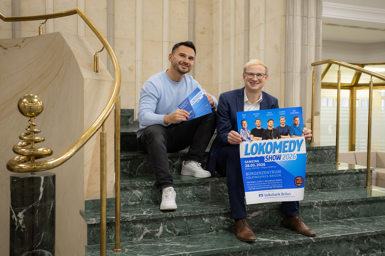 Zwei Männer sitzen auf einer geschwungenen Treppe und halten das Plakat für die „Lokomedy-Show“ 2026 in ihren Händen.