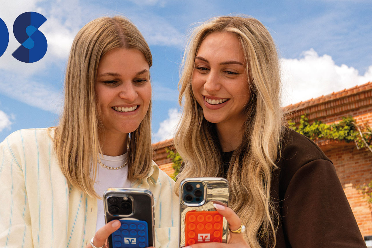 Zwei junge Frauen sitzen unter blauen Himmel und schauen lachend in ihre Handys