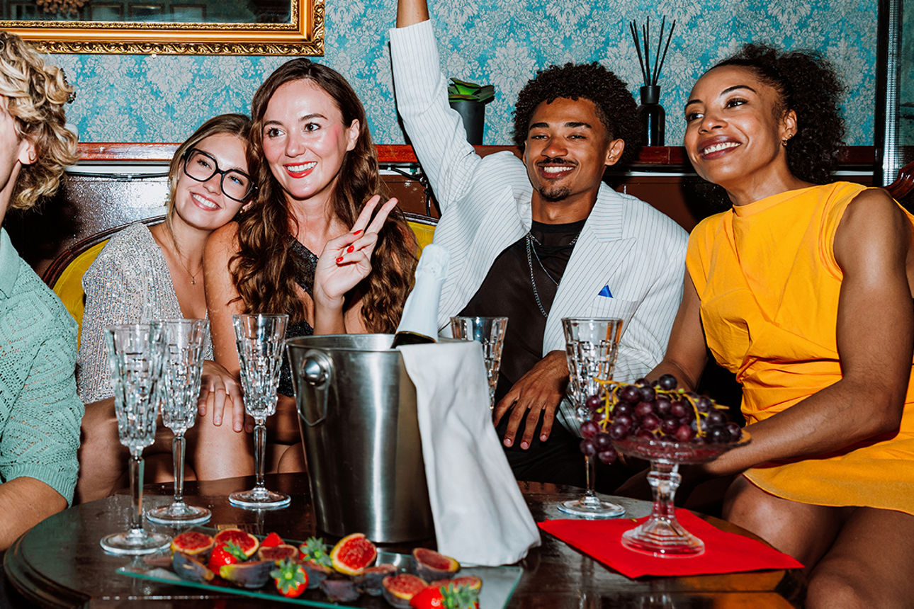 Gruppe von fünf Personen sitzt gemeinsam an einem Tisch in einer stilvollen Lounge und macht ein Selfie mit dem Smartphone.