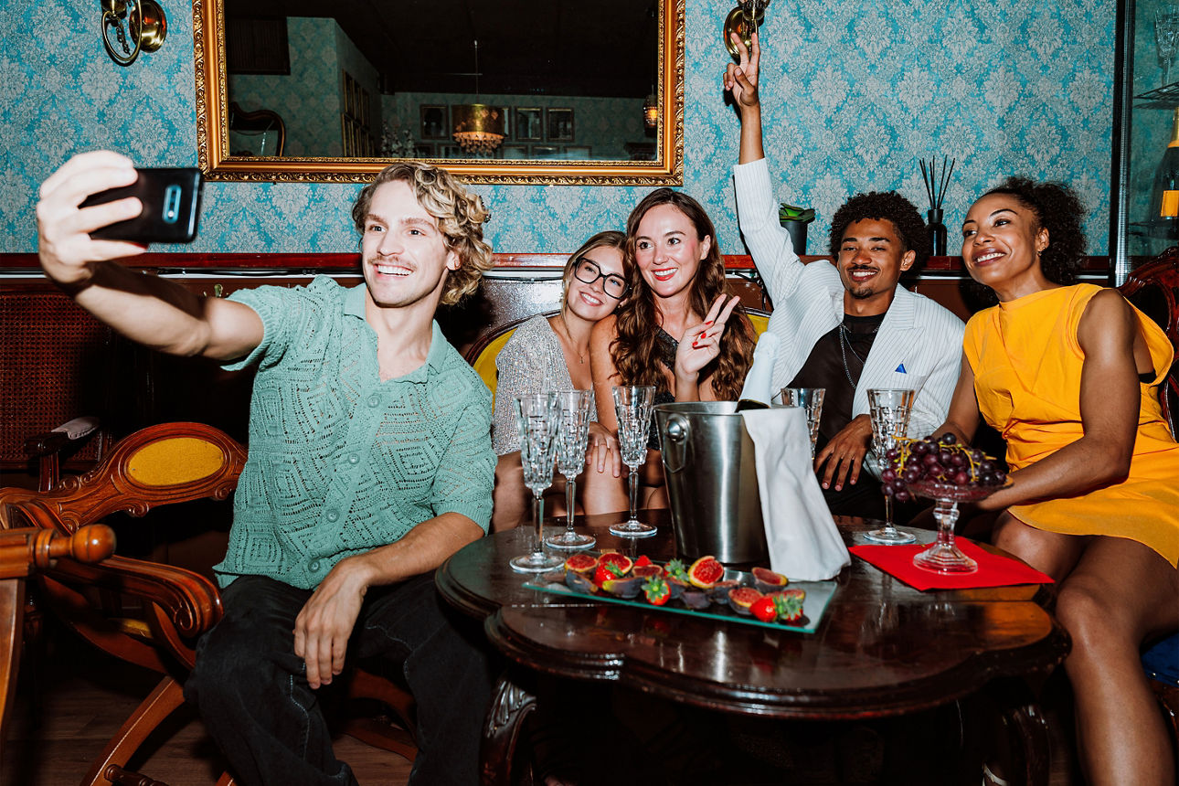 Gruppe von fünf Personen sitzt gemeinsam an einem Tisch in einer stilvollen Lounge und macht ein Selfi mit dem Smartphone.