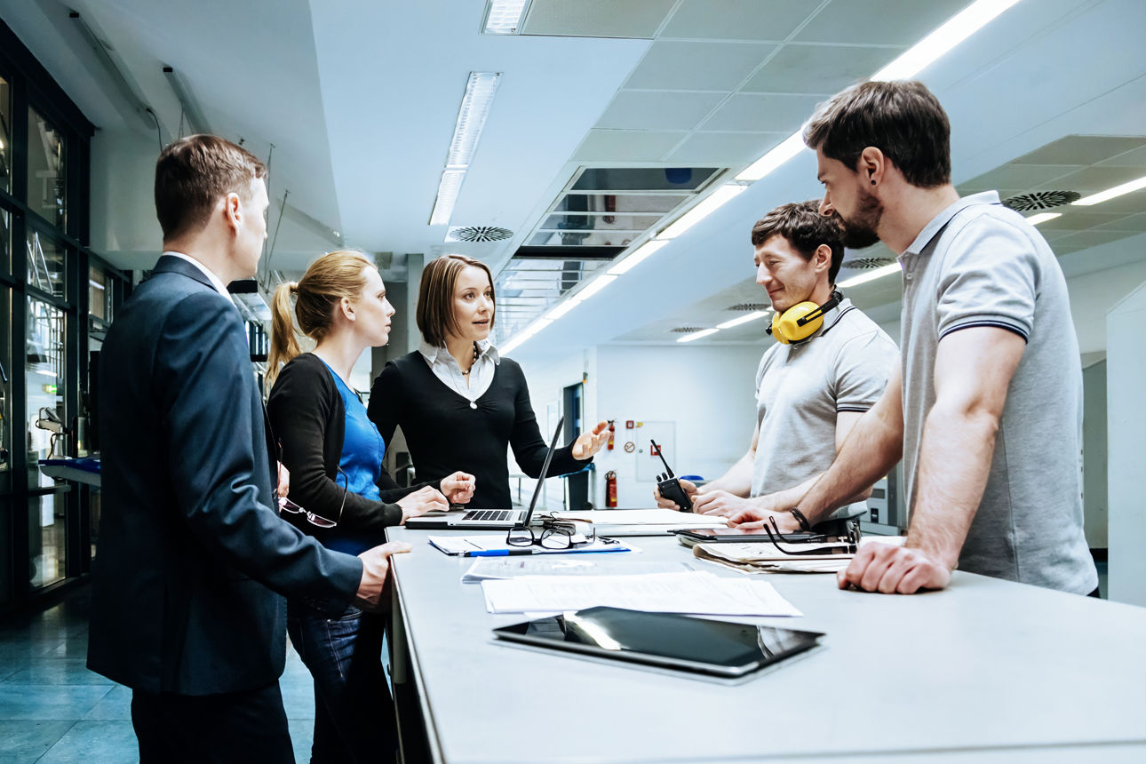 Fünf Personen bei einer Arbeitsbesprechung an einem Tisch in einer modernen Produktionshalle, auf dem ein Laptop, Tablets und Unterlagen liegen. Eine Frau im Business-Look spricht zur Gruppe, während Männer in Arbeitskleidung und Kolleginnen zuhören.
