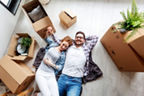 Aufsicht auf zwei Personen, die auf einer Decke zwischen mehreren Umzugskartons in einer hellen Wohnung liegen. Kartons mit Wohnaccessoires und Pflanzen stehen im Raum.