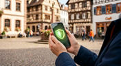 Zwei Hände halten schützend ein Smartphone mit einem grünen Schutzschild-Symbol auf dem Display vor dem unscharfen Hintergrund eines historischen Marktplatzes. Symbolfoto für das Zusatzpaket WIRSicher zum Schutz vor finanziellen Schäden durch Betrug (KI-generiert).