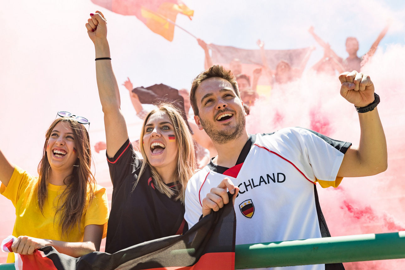3 Personen jublen bei einem Fußballspiel, eine Person im Deutschlandtrikot