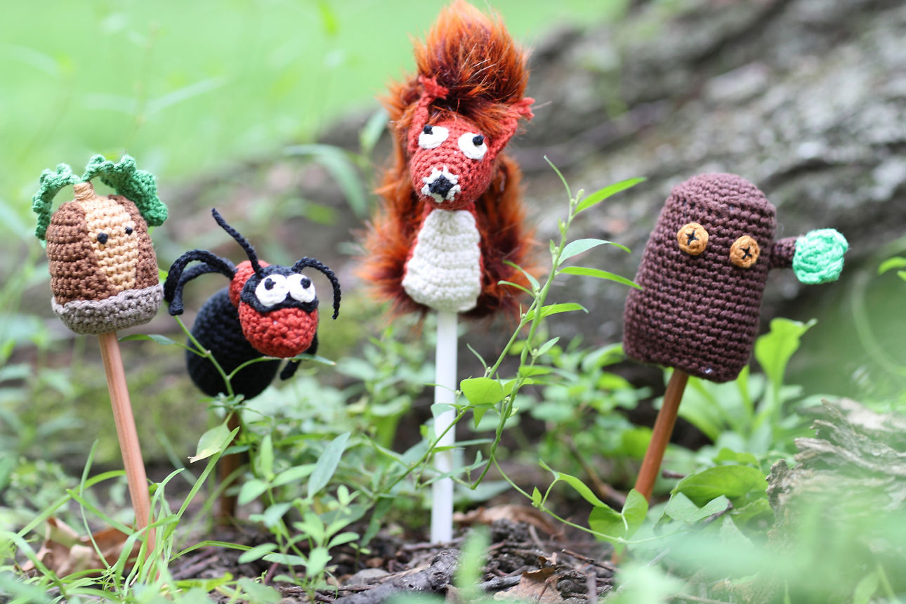 Eine Auswahl an liebevoll gehäkelten Wald- und Wiesen-Tieren steht auf einem Baumstamm im Freien. Zu sehen sind unter anderem ein Eichhörnchen, ein Vogel, eine Biene und verschiedene kleine Tiere und Pflanzenfiguren.