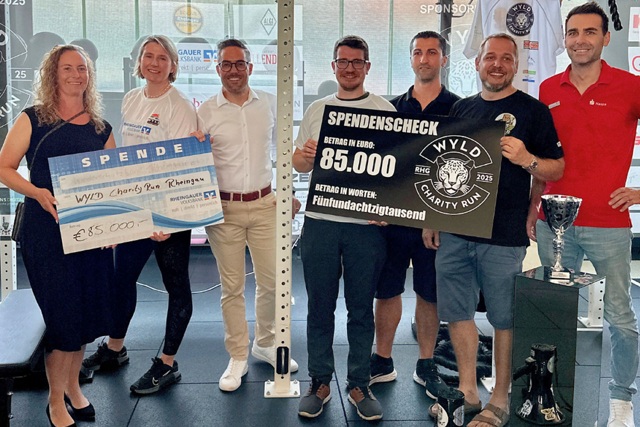 Gruppenszene mit fünf Personen, die einen großen Scheck mit 'SPENDENCHECK' und einen weiteren Scheck mit 'SPENDE' halten. Der Betrag auf dem Scheck ist '€85.000'. Im Hintergrund sind Fitnessgeräte und Sponsorenlogos sichtbar.