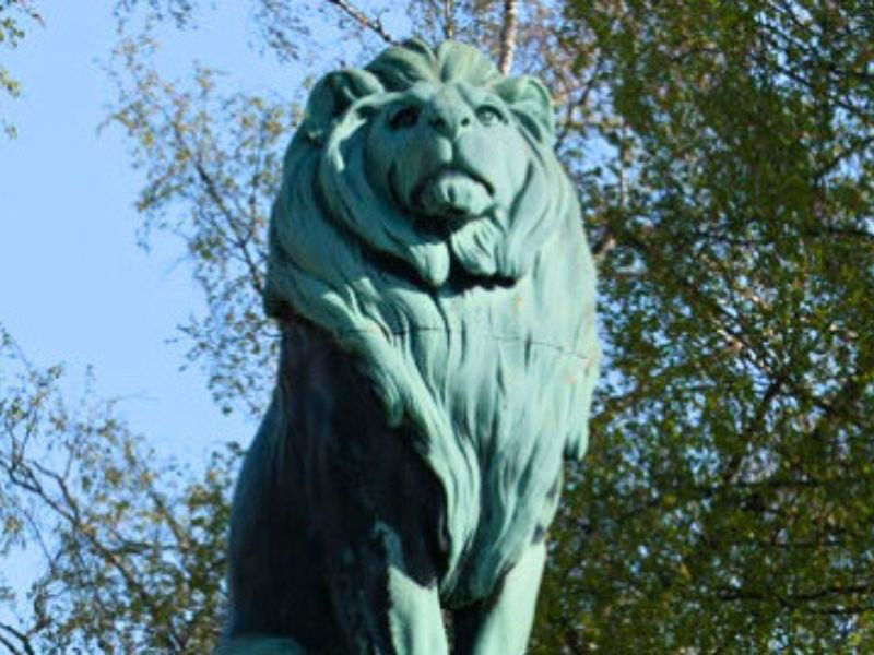 Eine beeindruckende Löwenstatue aus Bronze, umgeben von dem oberen Teil von Bäumen mit grünen und gelblichen Blättern unter einem strahlend blauen Himmel.