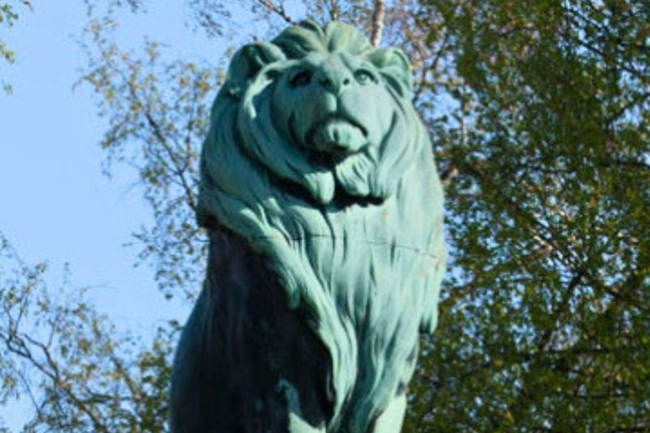 Eine beeindruckende Löwenstatue aus Bronze, umgeben von dem oberen Teil von Bäumen mit grünen und gelblichen Blättern unter einem strahlend blauen Himmel.
