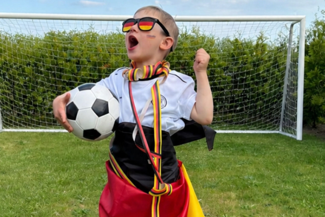 Ein Kind mit Deutschland‑Accessoires hält einen Fußball und steht jubelnd vor einem Tor im Garten.
