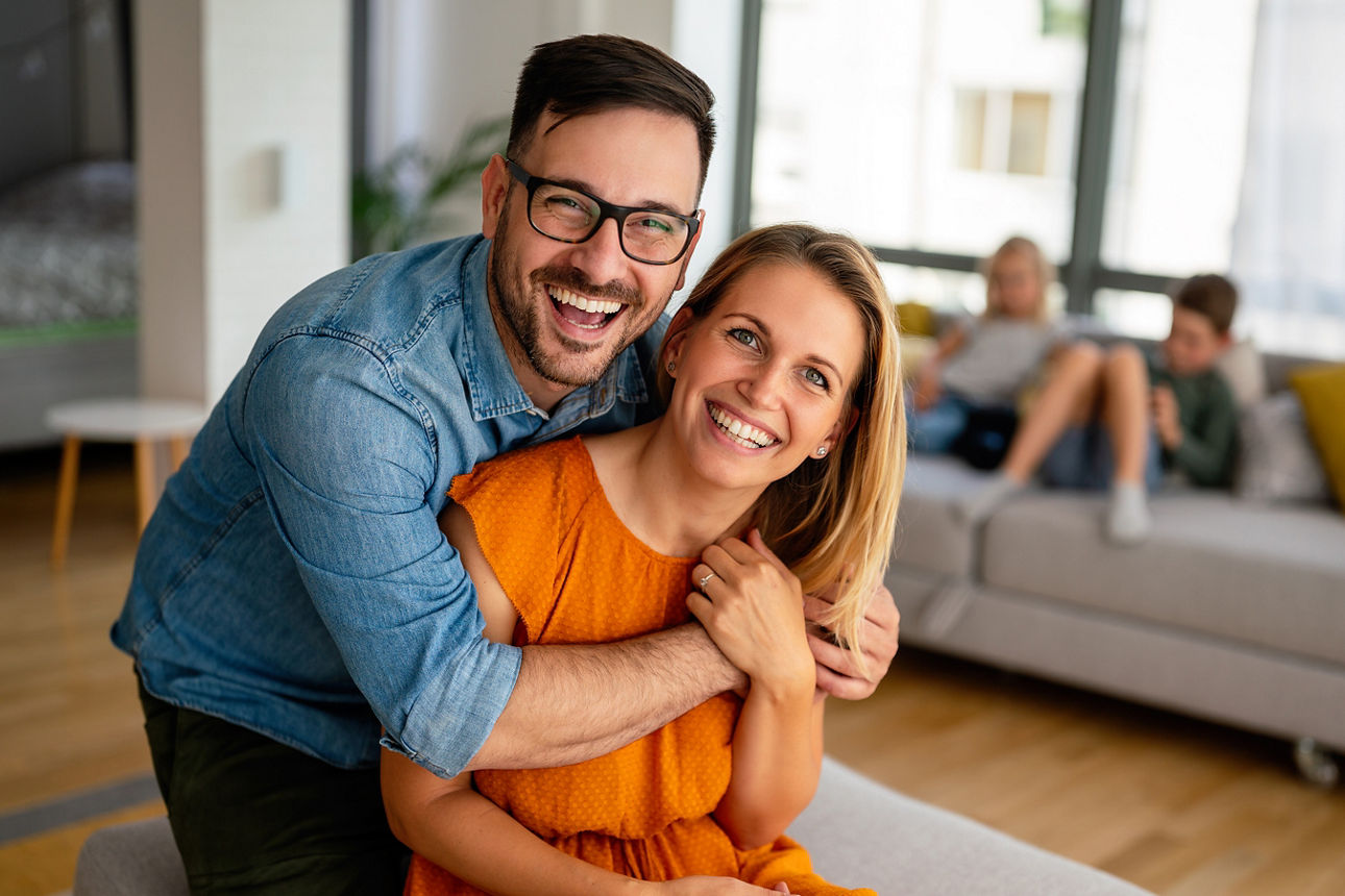 Mann in Jeanshemd umarmt Frau in orangefarbenem T-Shirt in einem Wohnzimmer mit Sofa und Kissen im Hintergrund.