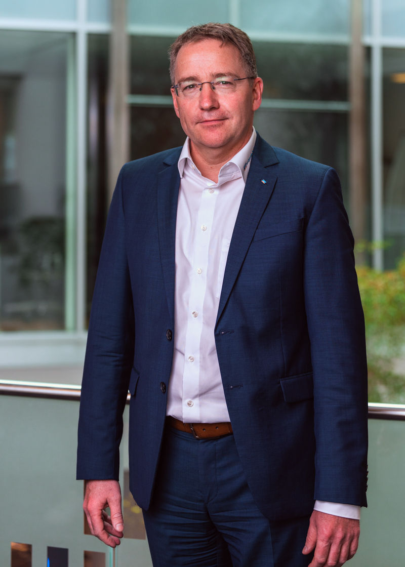 Ein Mann steht im dunklen Anzug vor einer Glaswand und lächelt in die Kamera.