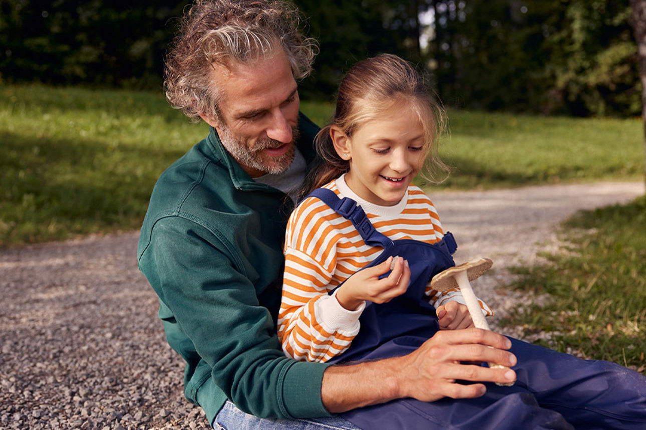 Mann sitzt mit Tochter am Wegesrand und betrachtet einen Pilz