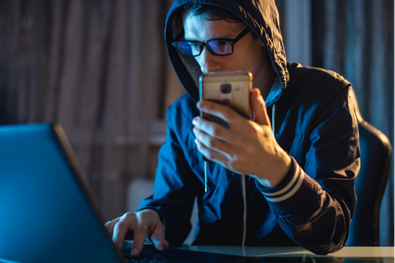 Hacker hält ein Telefon in der Hand und versucht mittels seines Notebooks Daten abzugreifen.