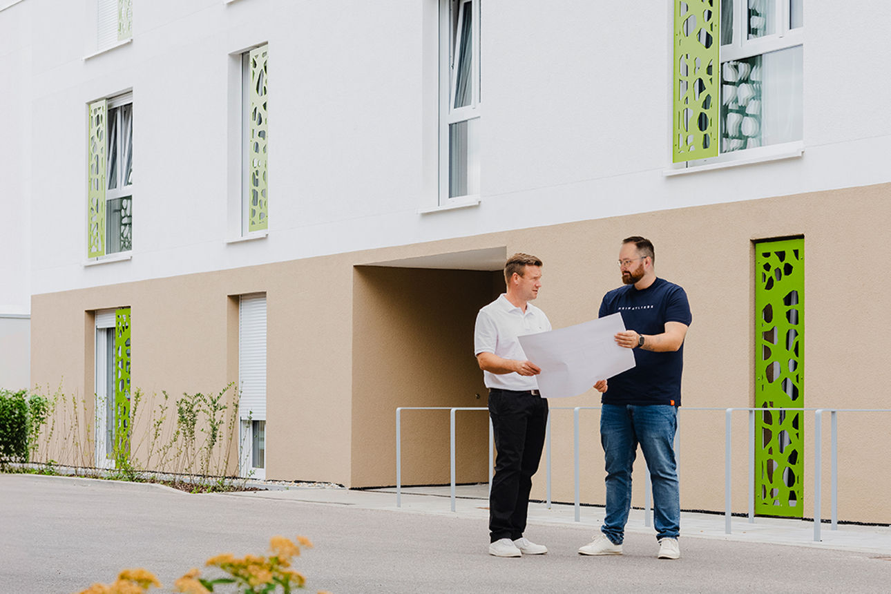 Zwei Personen stehen vor einem modernen Gebäude und betrachten gemeinsam einen Bauplan.