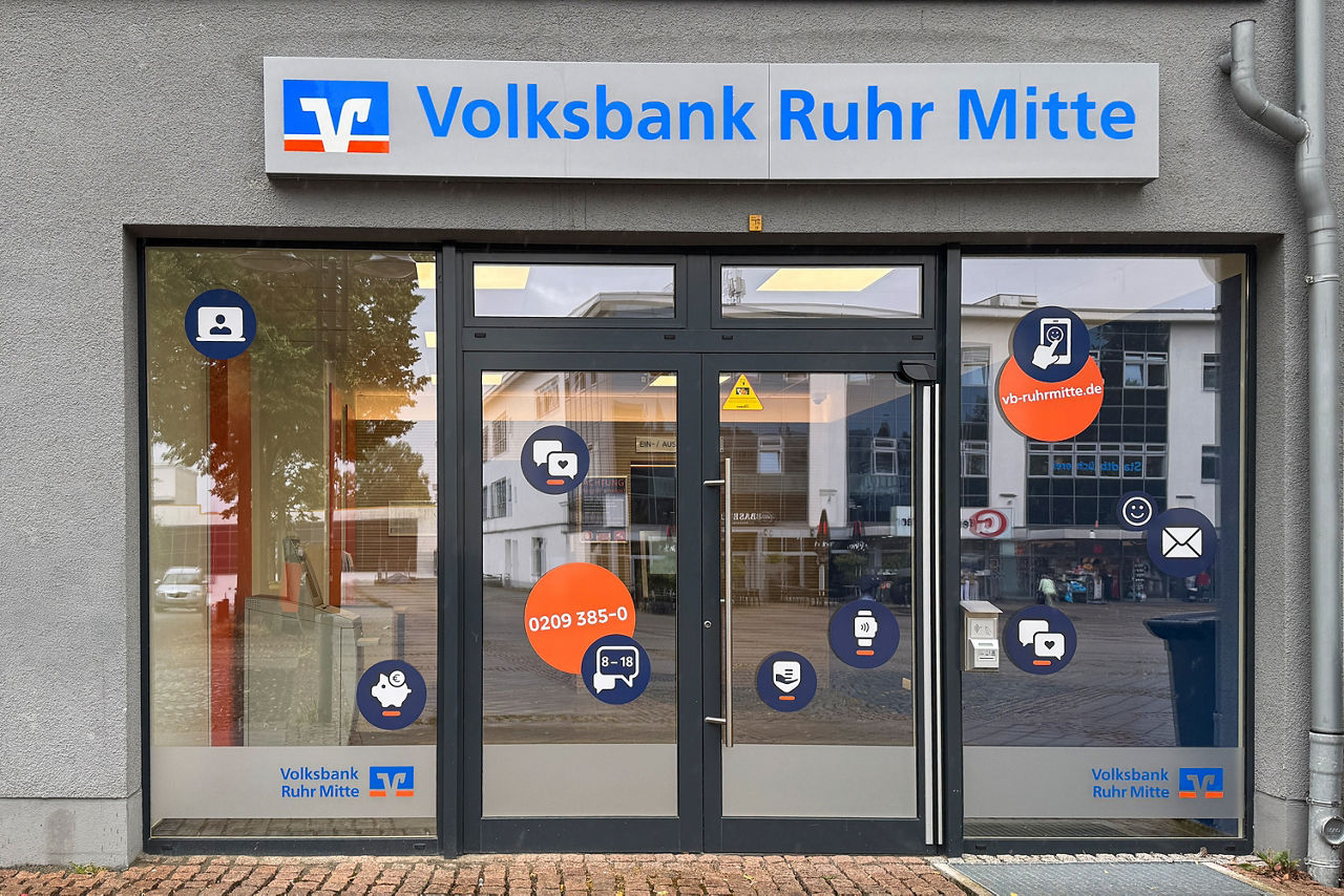 Zu sehen ist das SB-Centers Wattenscheid am Standort Alter Markt 8 von außen. Neben einer Doppelglastür ist rechts und links jeweils noch ein Fensterelement. Die Fensterfläche ist beklebt mit diversen Icons, die alle auf unterschiedliche Bankenthemen hinweisen. Es steht zudem die Telefonnummer und Internetadresse drauf.