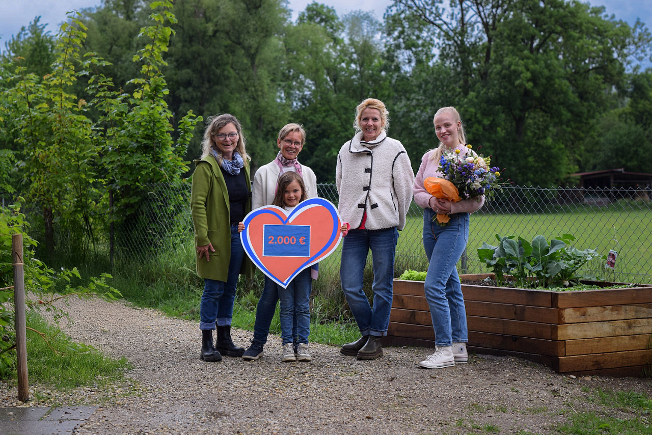 Vier Frauen und ein Mädchen in der Mitte mit Spendenherz in der Hand über 2.000 Euro stehen auf Kiesweg, die rechte Frau hält einen Blumenstrauß im Arm; im Hintergrund Wald und Wiese, rechts ein Hochbeet aus Holz