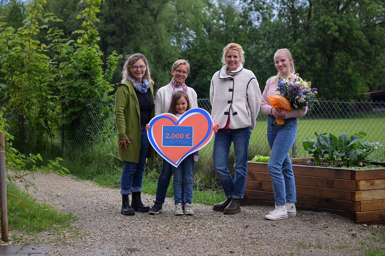 Vier Frauen und ein Mädchen in der Mitte mit Spendenherz in der Hand über 2.000 Euro stehen auf Kiesweg, die rechte Frau hält einen Blumenstrauß im Arm; im Hintergrund Wald und Wiese, rechts ein Hochbeet aus Holz