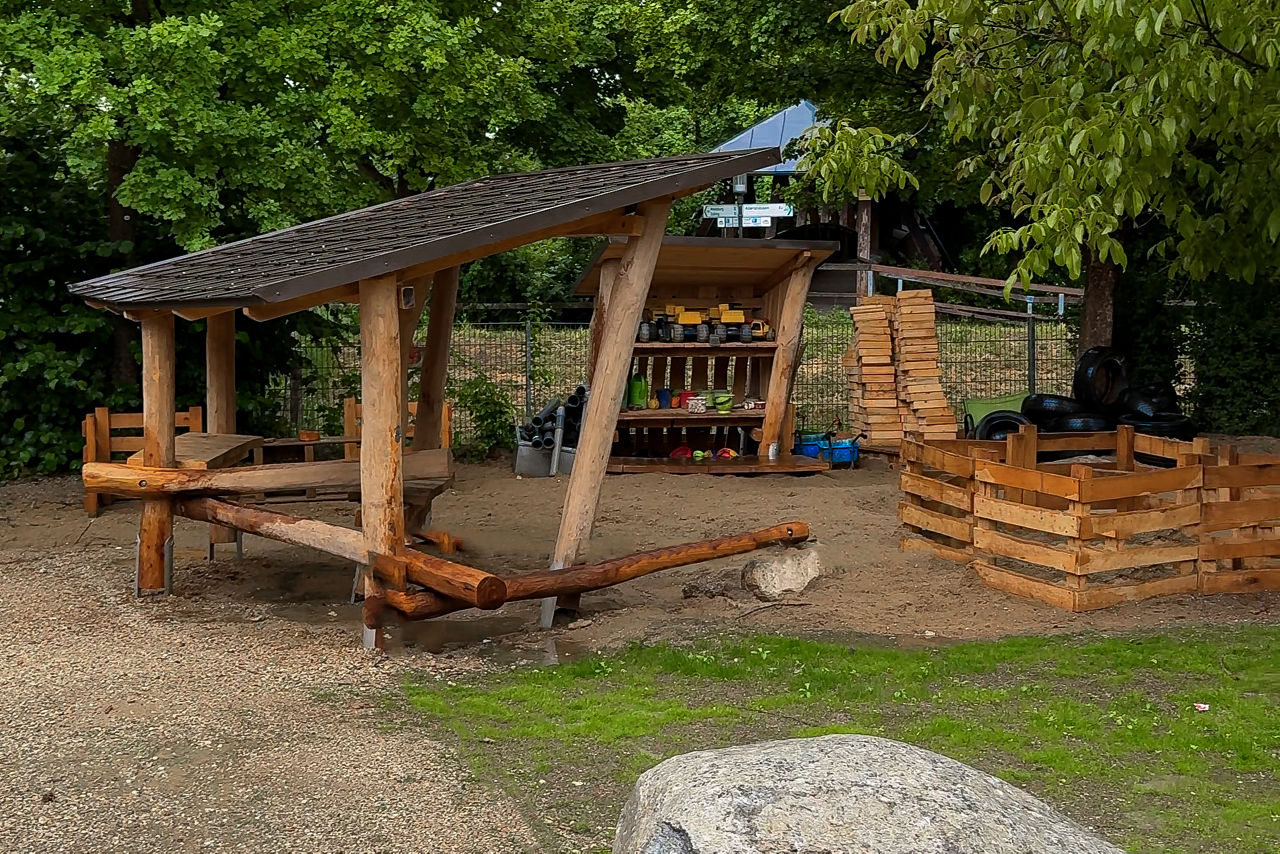 Spielplatz mit verschiedenen Holzbauten; links eine Überdachung mit dicken Stämmen als Gestell, rechts stehen Holzpaletten und aus Brettern gezimmerte Gestelle, für Hochbeete