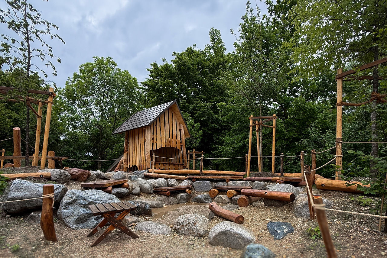 Kinderspielpaltz in der Natur mit einem Holzhaus mittig, davor ein Kreis aus großen Steinen und Holzstämmen als Sitz- und Klettermöglichkeiten