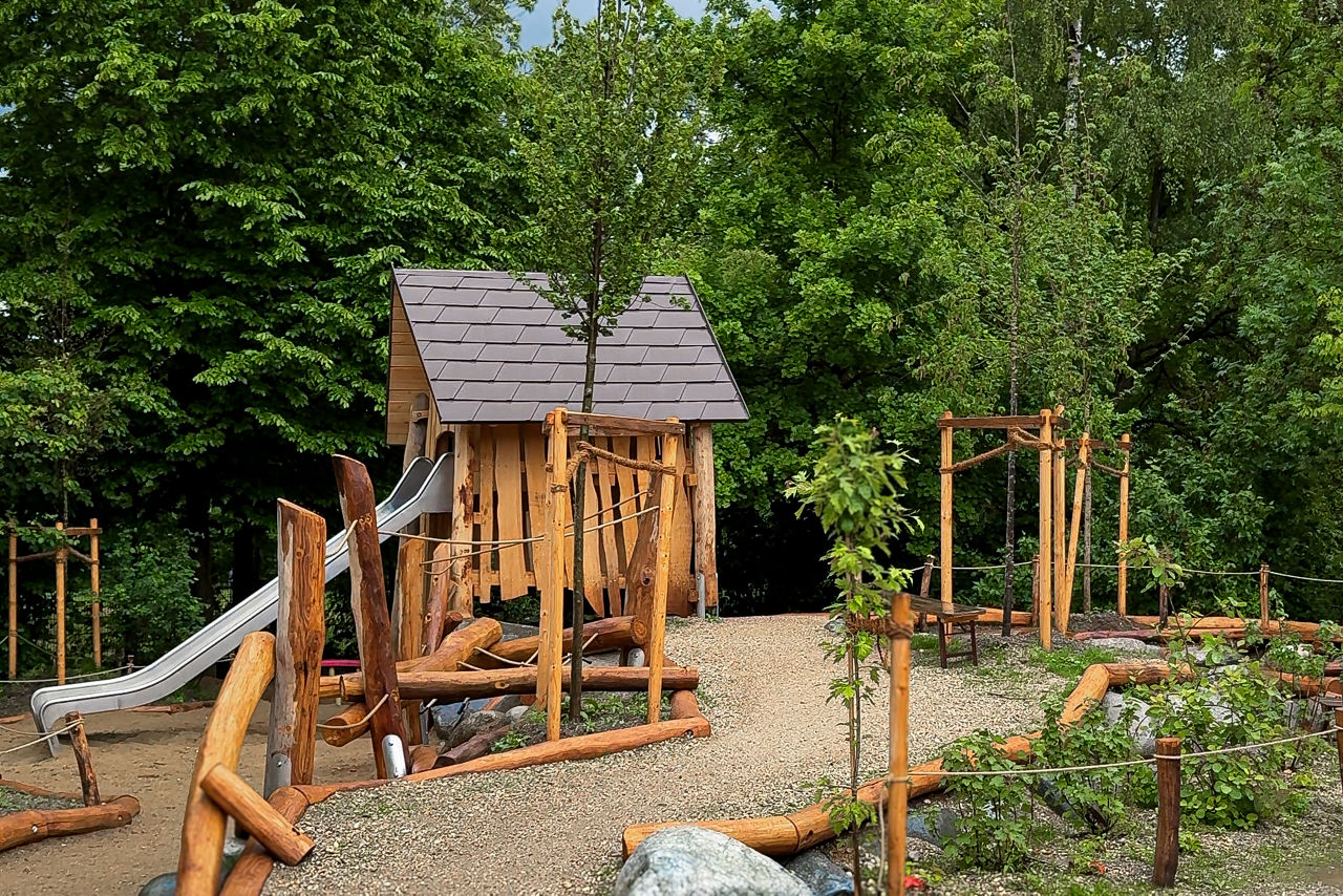 Holzhaus mit Rutsche vor dichtem Wald; Kiesweg führt geschwungen zwischen frischgepflanzten Bäumen durchs Bild