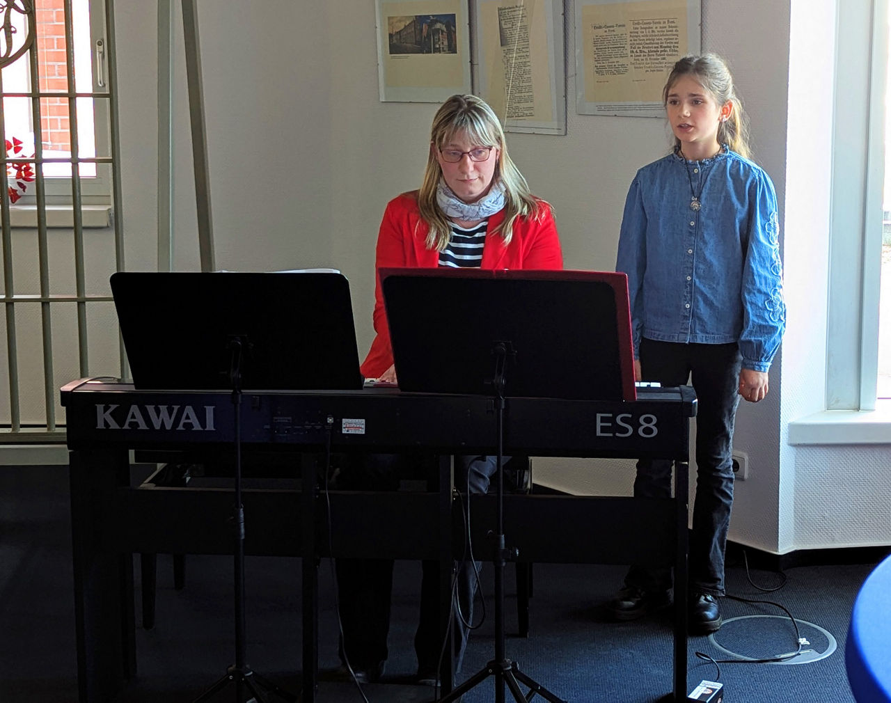 Eine Frau spielt am Klavier und ein Mädchen steht daneben und singt