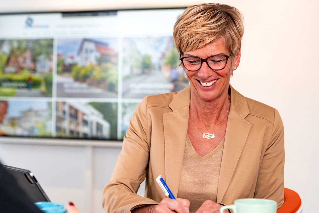 Ein Beratungsgesrpäch auf denen zwei Frauen zu sehen sind. Eine Frau lächelt und ist grade dabei etwas zu unterschreiben. 