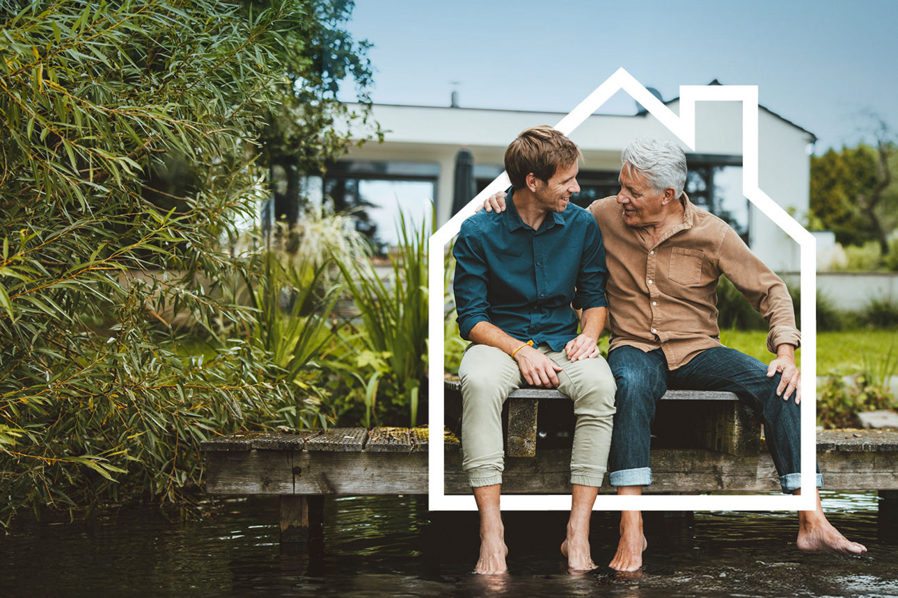 Junger und älterer Mann sitzen gemeinsam auf einem Holzsteg an einem Teich vor einem modernen Haus.