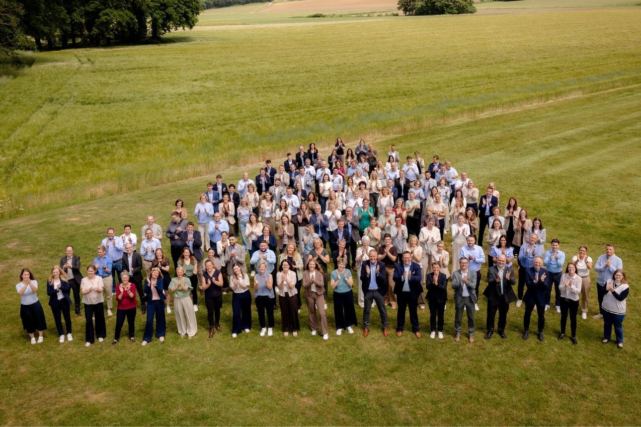 Mitarbeiter der Volksbank in der Hohen Mark eG stehen in mehreren Reihen auf einer Wiese im Freien und klatscht in Richtung Kamera; im Hintergrund offenes Grasland.