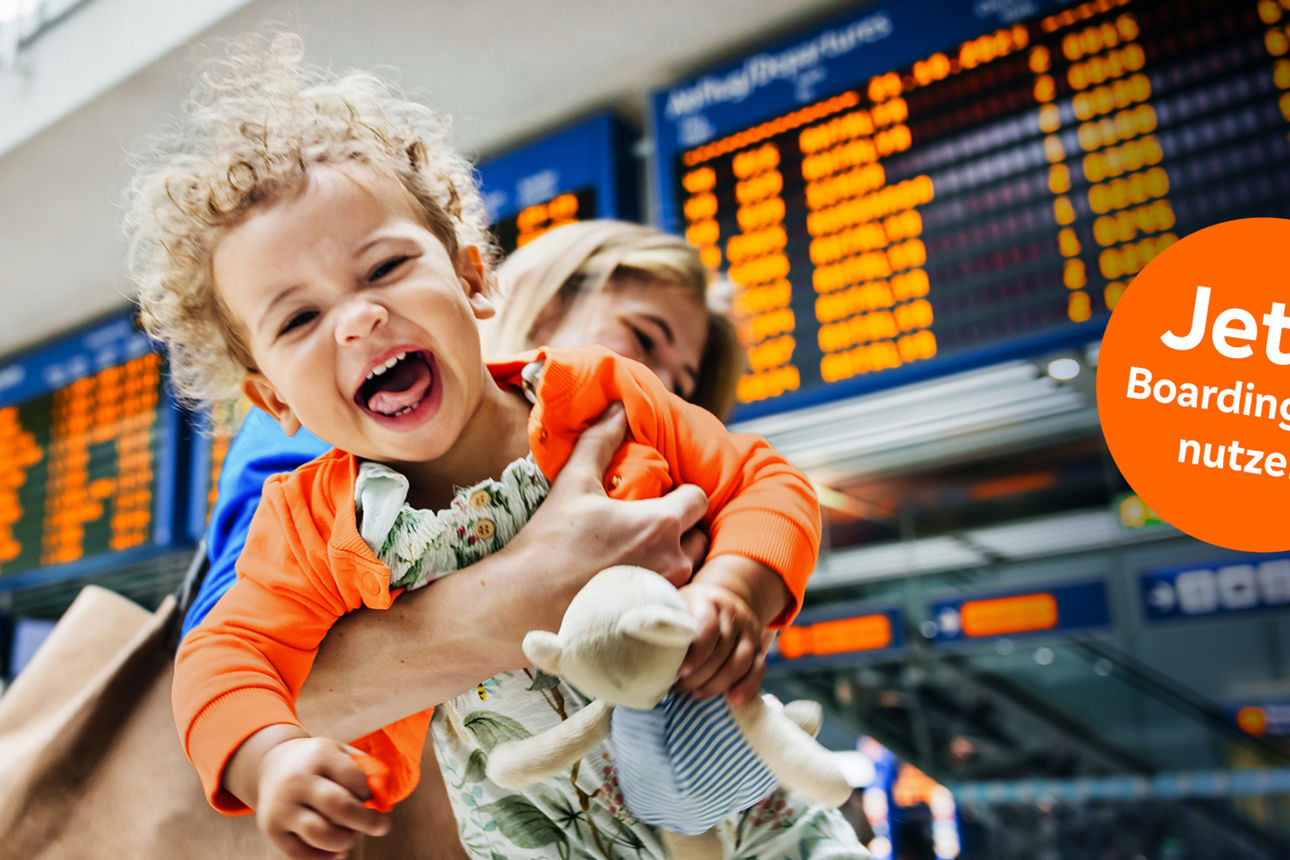 Junge Frau hält ihr lachendes Kind in den Armen, im Hintergrund eine Abflugtafel am Flughafen
