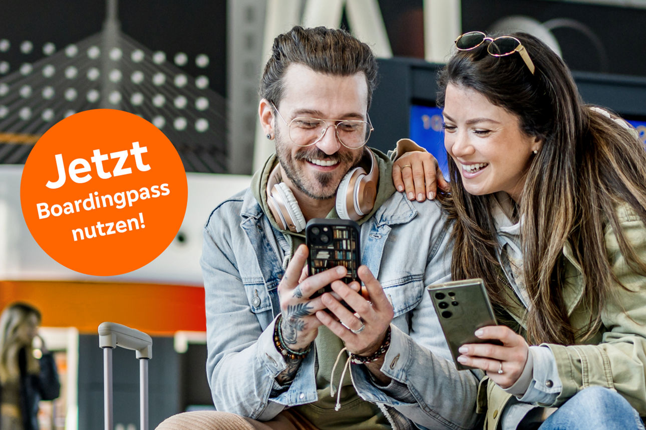 Pärchen sitzt mit Handy in der Hand und Koffern am Flughafen und lächelt