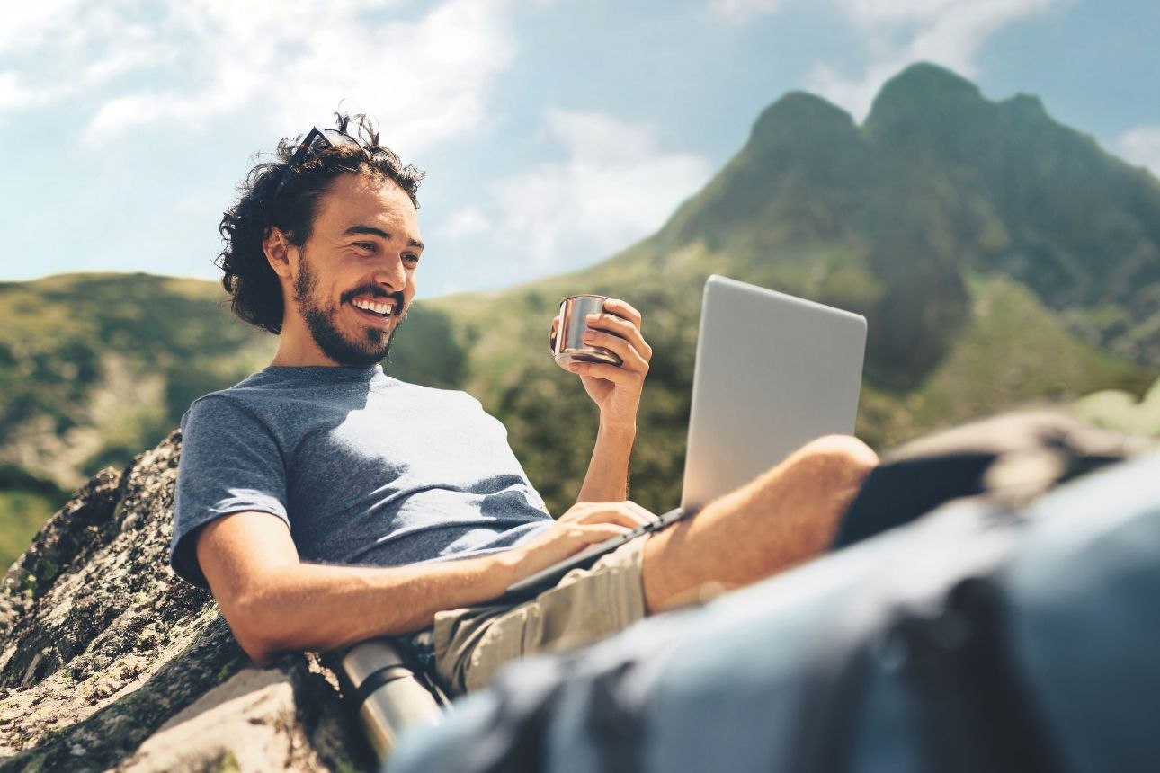 Person nutzt im Freien einen Laptop vor einer Berglandschaft und hält eine Tasse in der Hand.