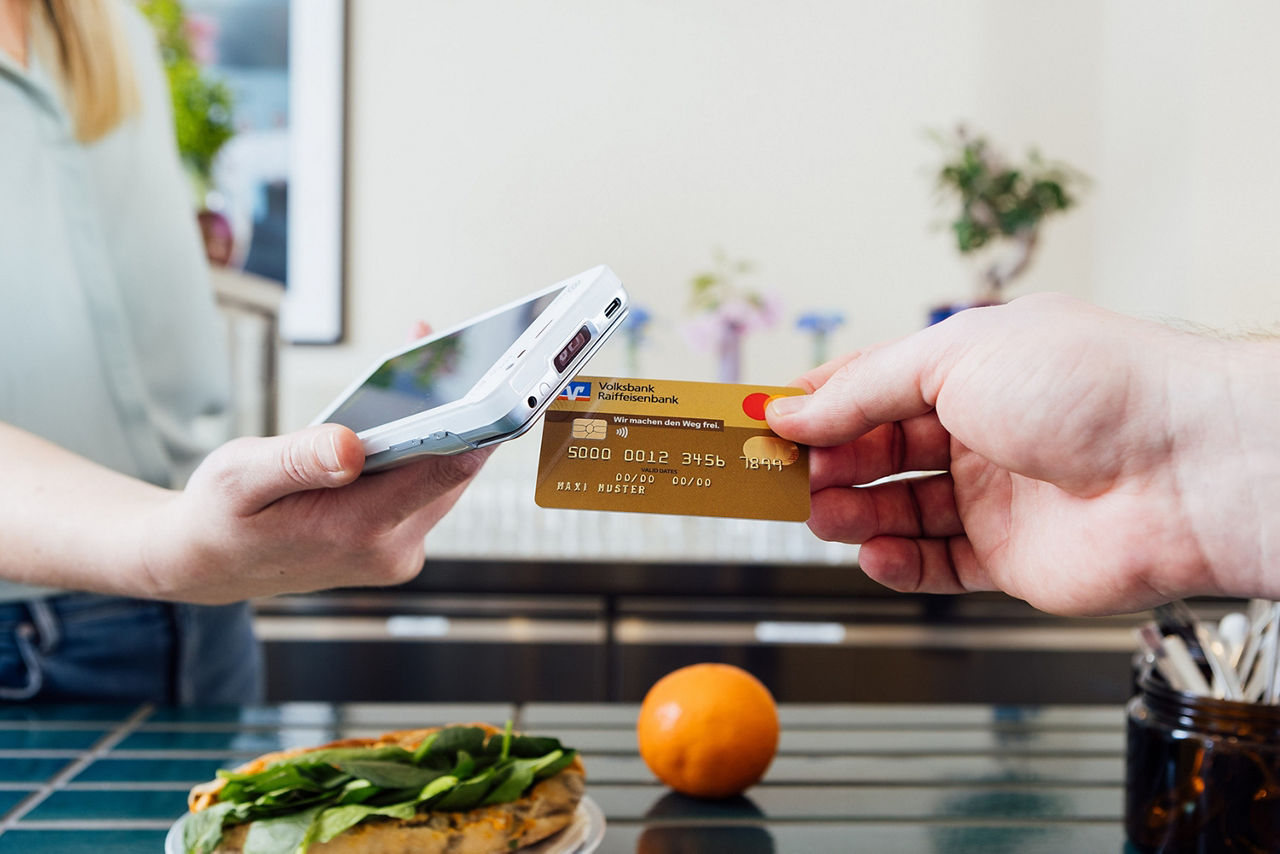 Eine blonde Frau reicht von links ein mobiles Kartenterminal ins Bild und von rechts hält die Hand einer weiteren Person eine goldene Kreditkarte an deses Terminal. 