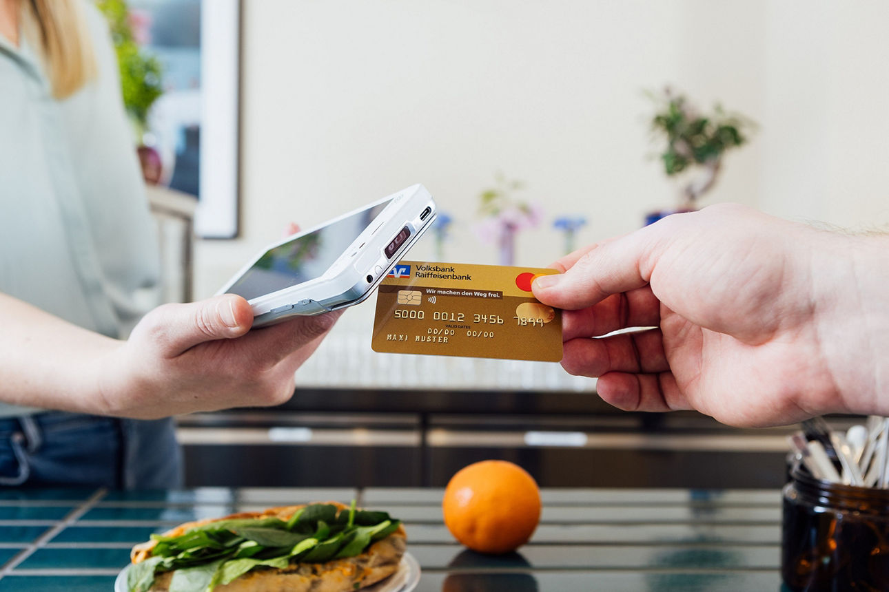 Eine blonde Frau reicht von links ein mobiles Kartenterminal ins Bild und von rechts hält die Hand einer weiteren Person eine goldene Kreditkarte an deses Terminal. 