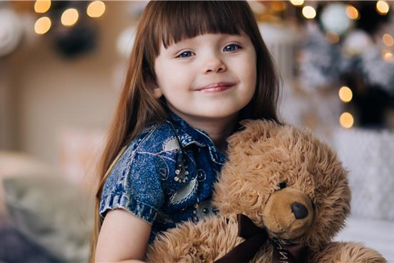 Ein braunhaariges Mädchen hat einen Teddy im Arm und lächelt in die Kamera.