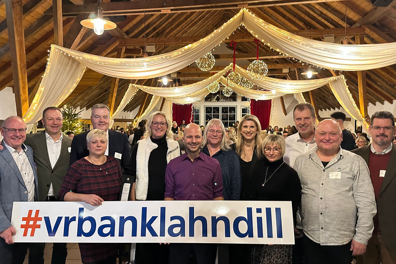 Gruppe von Menschen in festlich dekoriertem Raum, die ein Schild mit der Aufschrift '#vrbanklahndill' halten.