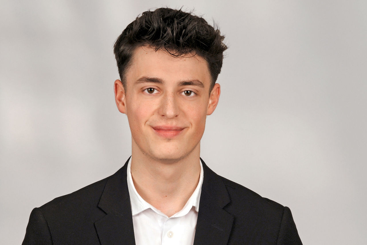 Junger Mann mit kurzen braunen Haaren trägt einen Anzug und lächelt in die Kamera