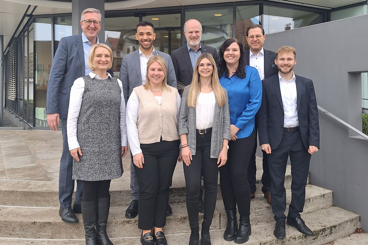 Auf dem Bild stehen vor einem Bankgebäude, auf einer Steintreppe, fünf Männer und vier Frauen und lächeln in die Kamera.