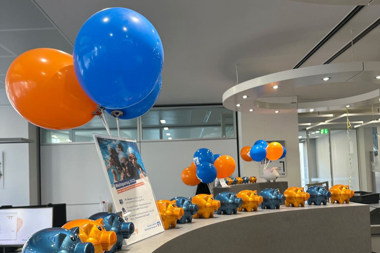 Blaue und orange Luftballons sowie Sparschweine auf einer Theke in einem modernen Büro.
